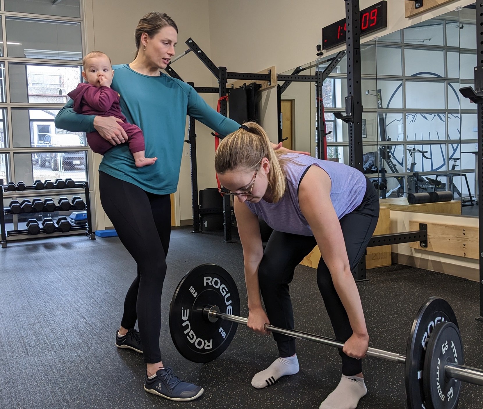 A pelvic floor physical therapist giving a patient cues for her deadlifting form while holding the patient's young baby, in Portland Maine.